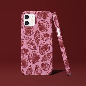 Könnte beinhalten: Eine rosa Handyhülle mit einem Muschelmuster. Die Hülle hat einen hellrosa Farbton mit einem sich wiederholenden Design aus dunkelroten Muscheln. Die Handyhülle steht aufrecht, mit einer passenden Hülle, die sich dagegen lehnt.