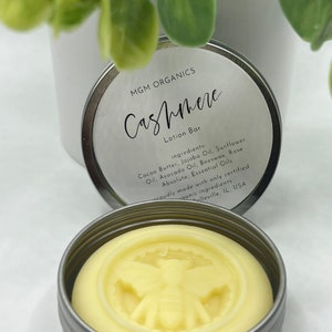 Może przedstawiać: Okrągły, jasnożółty balsam w srebrnej puszce. Na balsamie wytłoczony jest wzór pszczoły. Etykieta głosi "MGM ORGANICS Cashmere Lotion Bar" z listą składników. Produkt wykonany z organicznych składników w Belleville, IL, USA.