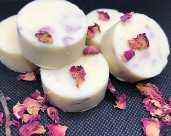 Puede incluir: Primer plano de varios jabones redondos de color blanquecino con pétalos de rosa secos incrustados. Los jabones están dispuestos sobre una superficie oscura, con pétalos de rosa esparcidos. Jabones artesanales para el cuidado de la piel.
