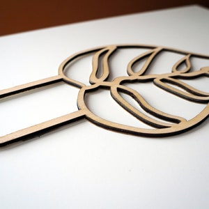May include: A wooden leaf-shaped cake topper with two pointed stakes. The topper is made of light-colored wood with a dark brown outline, and features a detailed leaf design. Perfect for adding a natural touch to cakes and desserts.