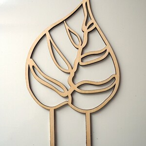May include: A wooden leaf-shaped garden stake with a cut-out design. The stake features a detailed leaf pattern with two pointed prongs at the base for easy insertion into soil. The natural wood color adds a rustic touch to any garden.