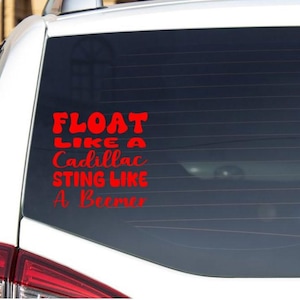 May include: A red vinyl decal on a car's rear window. The decal reads "FLOAT LIKE A Cadillac, STING LIKE A Beemer" in a stylized font. The car is white, and the window is tinted.