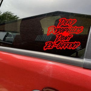 May include: A red car window with a red sticker that reads "Dead Pedophiles Don't Re-Offend?" in a stylized font. The sticker is placed on the window of a red vehicle, with the interior visible through the glass.