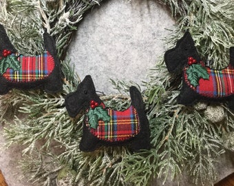 Könnte beinhalten: Drei schwarze Scottie-Hunde-Ornamente mit rot, grün und gelb karierten Satteldecken und Stechpalmenakzenten. Die Ornamente sind auf einem gefrosteten Tannenkranz angeordnet und schaffen eine festliche Weihnachtsdekoration.