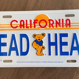 May include: White metal license plate with the text "CALIFORNIA DEADHEAD" in blue and red lettering. A cartoon bear with a blue bandana is in the center of the plate.