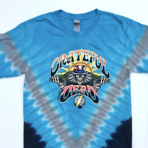 May include: A tie-dye t-shirt with a blue, gray, and black color scheme. The shirt features a graphic of the Grateful Dead band logo with a skeleton figure riding a motorcycle. The text "Grateful Dead" is written in a colorful, stylized font.