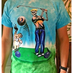 May include: A tie-dye t-shirt featuring a skeleton golfer with a golf club and a golf ball. The skeleton is wearing a yellow hat, a yellow shirt, blue jeans, and white shoes. A teddy bear with a golf club is standing on the green grass. The t-shirt has the text "GD TOUR" on the golf balls.
