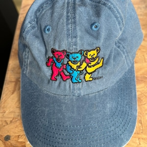 Pode incluir: Um boné de beisebol de denim azul com uma imagem bordada de três ursos dançando. Os ursos são vermelho, amarelo e azul. O boné tem uma aba curva e uma correia ajustável azul.
