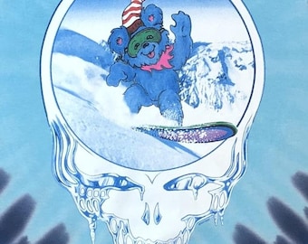 May include: A tie-dye blue and black t-shirt featuring a white outline of a smiling skull with dripping icicles. Inside the skull is a blue bear wearing a red and white hat and goggles, snowboarding down a snowy mountain.