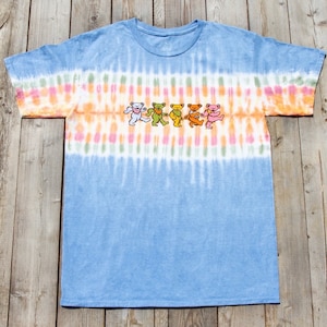 Puede incluir: Una camiseta azul claro tie-dye con una banda de tie-dye de colores a través del pecho. La camiseta presenta un gráfico de cinco osos bailando en línea.