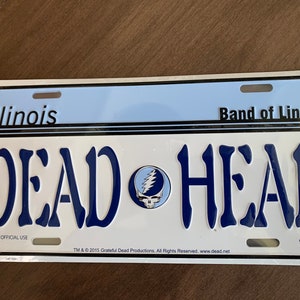 Może przedstawiać: Biała metalowa tablica rejestracyjna z niebieskim tekstem, który brzmi "Illinois Band of Lincoln DEAD HEAD" z niebiesko-białym logo Grateful Dead w środku. Dolna część tablicy zawiera napis "NOT FOR OFFICIAL USE" i "TM & © 2015 Grateful Dead Productions. All Rights Reserved. www.dead.net" mniejszym tekstem.