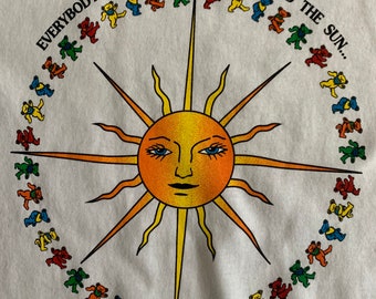 Puede incluir: Una camiseta blanca con un gráfico de un sol amarillo con una cara y un círculo de osos danzantes de colores alrededor. El texto "Everybody's dancin' in a ring around the sun..." está impreso alrededor de los osos.