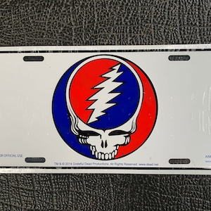 May include: White metal license plate featuring a colorful Grateful Dead Steal Your Face logo. The logo is a red, white, and blue skull with a lightning bolt. The text "NOT FOR OFFICIAL USE" and "AIMGIFTS MADE IN USA" are printed on the plate.
