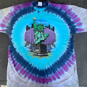 May include: Tie-dye t-shirt with a blue, green, and purple swirl pattern. The shirt features a cartoon image of the Statue of Liberty with the words "NEW YORK" in a colorful font.
