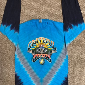 May include: A blue tie-dye long-sleeve t-shirt with a Grateful Dead graphic featuring a skeleton wearing a top hat and sunglasses, riding a motorcycle. The graphic also includes the words "Grateful Dead" and a lightning bolt.