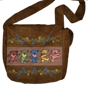 May include: Brown corduroy shoulder bag with an embroidered design. The design features dancing bears in various colors, with blue and yellow floral accents. The bag has a long strap for over-the-shoulder or crossbody wear.