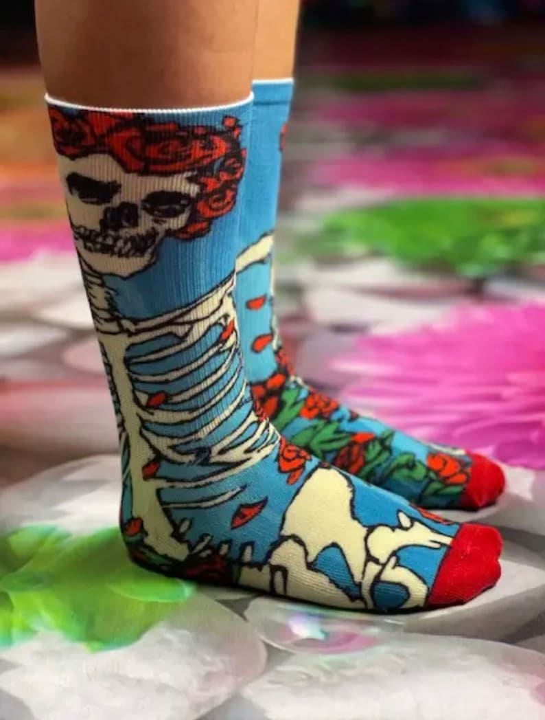 K&ouml;nnte beinhalten: Ein Paar blaue Socken mit einem Totenkopf- und Skelett-Design. Der Totenkopf tr&auml;gt eine Krone aus roten Rosen. Die Socken haben einen roten Saum oben und unten.