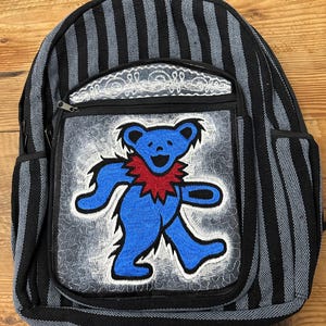 May include: Black and gray striped backpack with a blue dancing bear patch. The bear has a red collar and is outlined in white. The backpack has a front pocket with a decorative lace trim and two side pockets.
