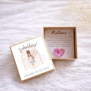 Könnte beinhalten: Eine kleine, offene, quadratische Geschenkbox mit einer Aquarellillustration eines Mädchens, das einen Blumenstrauß und einen Regenbogen hält. Der Deckel der Box trägt die Aufschrift "Schulkind" und "Johanna 28.08.2026". Innen ein rosa herzförmiger Anhänger.