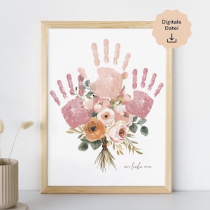 Op de afbeelding: Een ingelijste print met roze en perzik handafdrukken rond een bloemenboeket. Het boeket bevat oranje en roze rozen. De tekst "MIT liebe VON" staat onderaan, met een digitaal downloadpictogram rechtsboven.