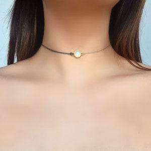 May include: A delicate choker necklace featuring a small, round, iridescent gemstone pendant. The necklace is made of thin, black cord and has a simple, minimalist design.