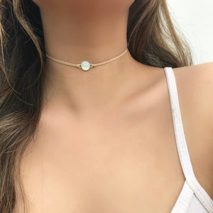 Può includere: Una delicata collana girocollo con un cordoncino beige chiaro e un piccolo pendente rotondo con una pietra opalescente. La collana è indossata intorno al collo, evidenziando il suo design minimalista e la sua sottile eleganza.
