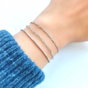 Puede incluir: Tres pulseras de cadena plateadas apiladas en una mu&ntilde;eca. La pulsera superior es una cadena fina y delicada. La pulsera del medio es una cadena m&aacute;s gruesa con un dise&ntilde;o de eslabones. La pulsera inferior es una cadena de eslabones cuadrados.