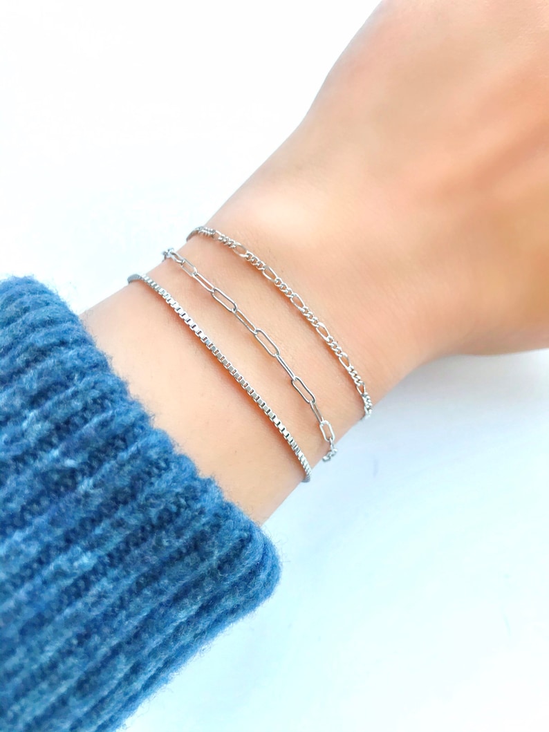 Puede incluir: Tres pulseras de cadena plateadas apiladas en una mu&ntilde;eca. La pulsera superior es una cadena fina y delicada. La pulsera del medio es una cadena m&aacute;s gruesa con un dise&ntilde;o de eslabones. La pulsera inferior es una cadena de eslabones cuadrados.