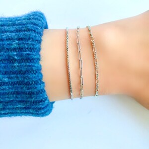 Puede incluir: Tres pulseras de cadena plateadas apiladas en una mu&ntilde;eca. La pulsera superior es una cadena fina y delicada. La pulsera del medio es una cadena m&aacute;s gruesa con eslabones ovalados. La pulsera inferior es una cadena m&aacute;s gruesa con eslabones cuadrados.