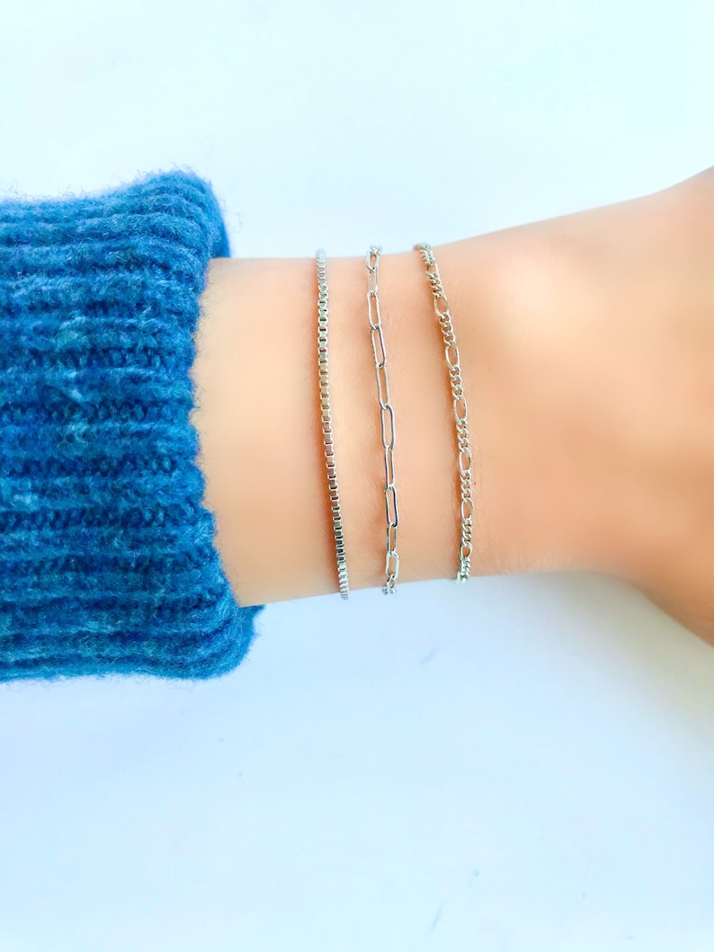 Puede incluir: Tres pulseras de cadena plateadas apiladas en una mu&ntilde;eca. La pulsera superior es una cadena fina y delicada. La pulsera del medio es una cadena m&aacute;s gruesa con eslabones ovalados. La pulsera inferior es una cadena m&aacute;s gruesa con eslabones cuadrados.