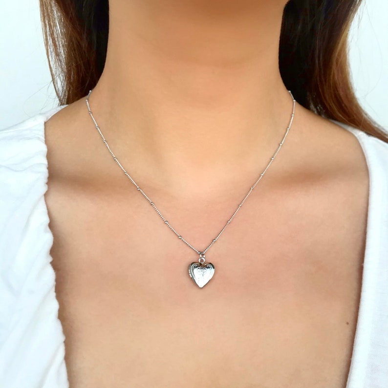 May include: A silver heart-shaped locket necklace on a delicate chain. The locket has a detailed design and is a classic piece of jewelry.