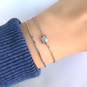 Puede incluir: Dos pulseras de plata en una muñeca. La pulsera superior tiene un pequeño dije de tortuga.