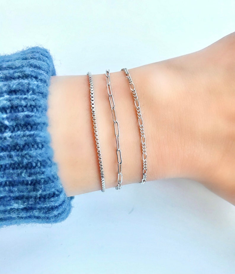 Puede incluir: Tres pulseras de cadena plateadas en una mu&ntilde;eca. Las pulseras son de diferentes estilos, incluyendo una cadena de caja, una cadena de clip de papel y una cadena de bordillo.