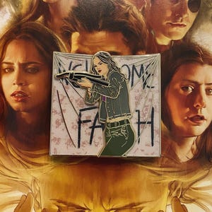 May include: Enamel pin depicting a woman with a crossbow, with the text "WELCOME FAITH" on a banner. The woman wears a green jacket and trousers. The background shows a collage of faces.