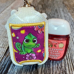 May include: A white glitter hand sanitizer holder with a purple and gold embroidered design featuring a green chameleon. The holder is attached to a silver key chain. The holder is filled with a small bottle of PocketBac hand sanitizer with a red label that says "Champagne Apple Honey" and "Anti-Bacterial Hand Gel with Essential Oils 1 fl oz/29 mL".