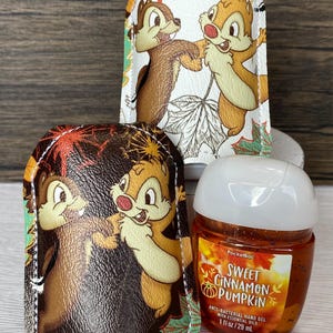May include: Three items are displayed: two hand sanitizer holders and a hand sanitizer bottle. The holders feature Chip and Dale cartoon characters. The hand sanitizer bottle is orange and has the text "Sweet Cinnamon Pumpkin".