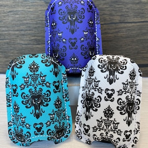 May include: Three small, rectangular pouches with rounded tops. Each pouch features a repeating black pattern on a colored background: blue, teal, and white. The pattern includes stylized figures and owl-like faces. White stitching outlines the edges.