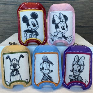 May include: Five different hand sanitizer holders with embroidered images of Disney characters. The holders are made of different colored leather and have a clear plastic window to view the sanitizer bottle. The characters are Mickey Mouse, Minnie Mouse, Pluto, Donald Duck, and Daisy Duck.