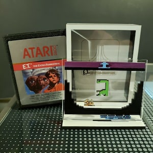 May include: An Atari E.T. video game cartridge and a display featuring pixelated game graphics. The cartridge has the game title and movie poster art. The display shows a green pixelated creature and the Atari logo.