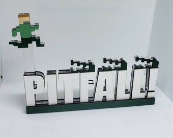 May include: A decorative sign featuring the word "PITFALL!" in white block letters with a green base. A pixelated figure in green and tan is jumping over a series of obstacles. The sign is made of wood and acrylic.