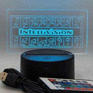Könnte beinhalten: Ein blaues LED-Licht mit einer durchsichtigen Acrylplatte, auf der das Intellivision-Logo und ein Raster aus pixelartigen Figuren abgebildet sind. Das Licht steht auf einem schwarzen Sockel mit einem USB-Stromkabel und einer Fernbedienung.