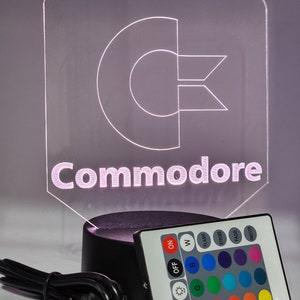 May include: A clear acrylic lamp featuring a white outline of the Commodore logo with the word "Commodore" underneath. The lamp sits on a black base and includes a remote control with buttons for on, off, flash, strobe, fade, smooth, and colour selection.