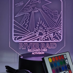 Puede incluir: Una lámpara de acrílico transparente con una base negra y un mando a distancia. La lámpara presenta un gráfico del clásico videojuego River Raid, con el texto "River Raid Released 1982 Activision". El mando a distancia tiene botones para diferentes colores de luz y efectos.