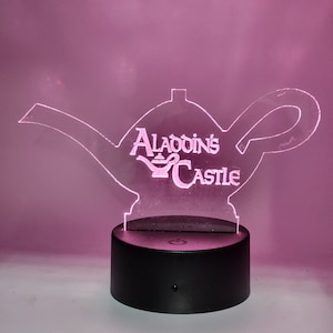 May include: A clear acrylic lamp shaped like a teapot with the words "Aladdin's Castle" etched on the side. The lamp is lit up with a pink light and sits on a black base with a power switch.