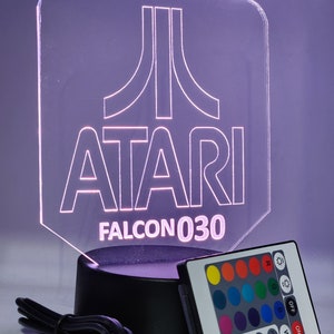 Puede incluir: Una lámpara de acrílico transparente con un contorno blanco del logotipo de Atari y el texto "ATARI" y "FALCON030". La lámpara tiene una base negra y viene con un mando a distancia con opciones de cambio de color.