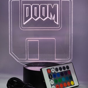 May include: A clear acrylic lamp shaped like a floppy disk with the word "DOOM" in white lettering. The lamp has a black base and comes with a remote control with multiple color options.