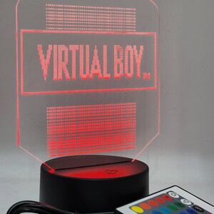 以下が含まれることがあります： 「VIRTUAL BOY」という白い文字が書かれた赤い照明付きアクリル看板。黒い台座に置かれており、USBケーブルと、複数の色のボタンが付いたリモコンが付属しています。