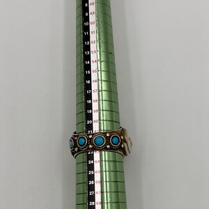 May include: A green metal ring sizer with a white and red numbered scale. The scale ranges from size 41 to 73. A silver ring with turquoise stones is placed on the sizer at size 61.