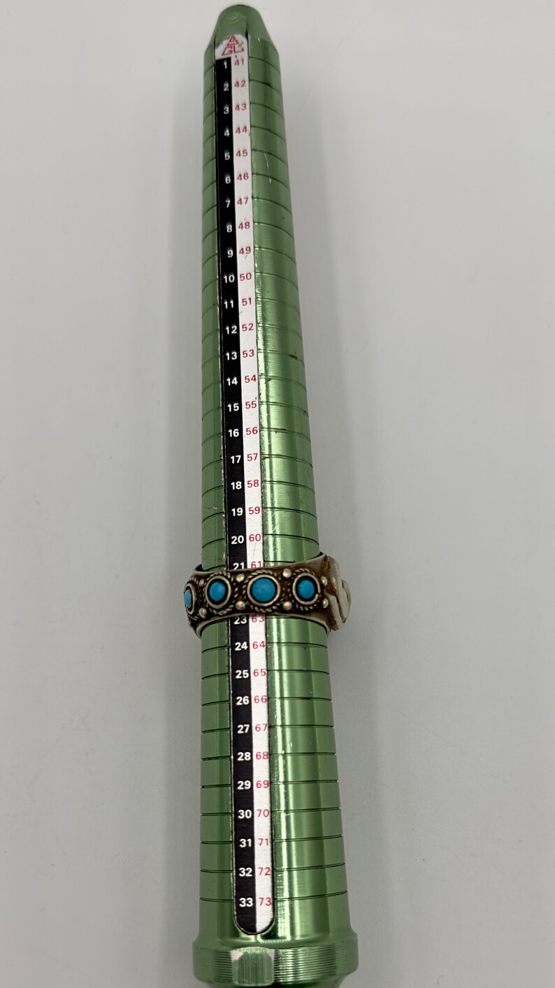 May include: A green metal ring sizer with a white and red numbered scale. The scale ranges from size 41 to 73. A silver ring with turquoise stones is placed on the sizer at size 61.