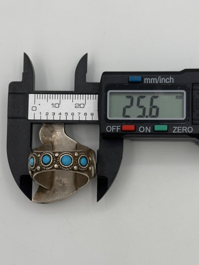 May include: A silver ring with turquoise stones and intricate detailing, measured at 25.6 mm in diameter.